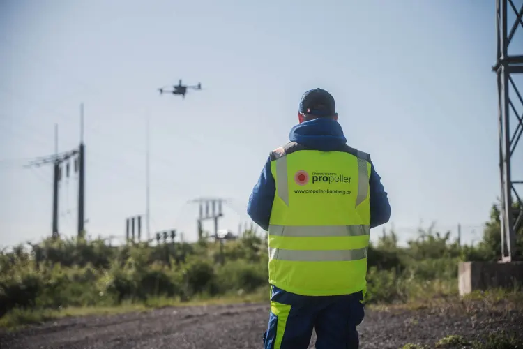 Pilot in Warnweste vor einem Umspannwerk, die Drohne fliegt im Hintergrund